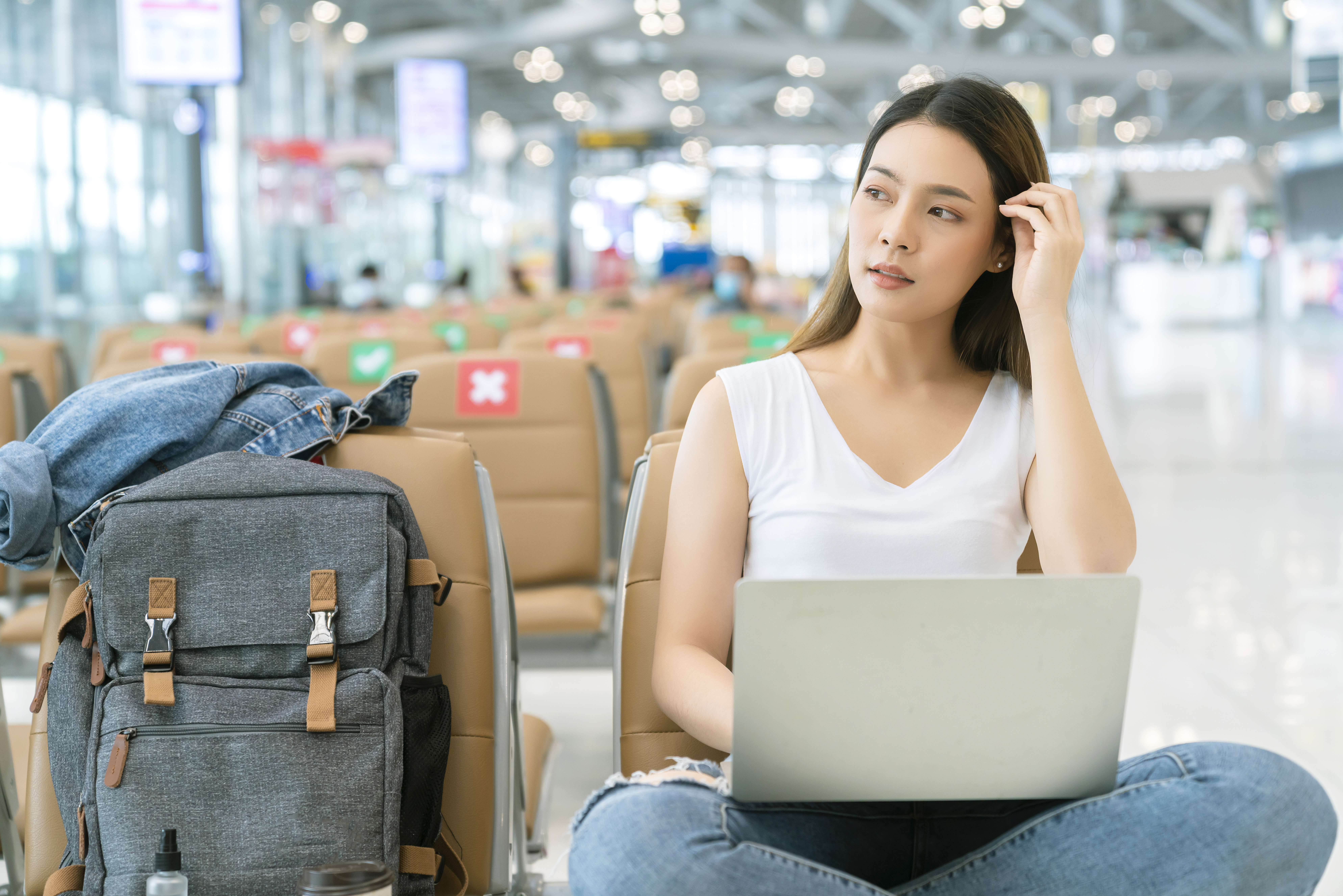 attractive-asian-female-casual-cloth-with-backpack-traveller-relax-sit-wait-social-distancing-seat-departure-airport-terminal-hand-use-laptop-safety-travel-concept