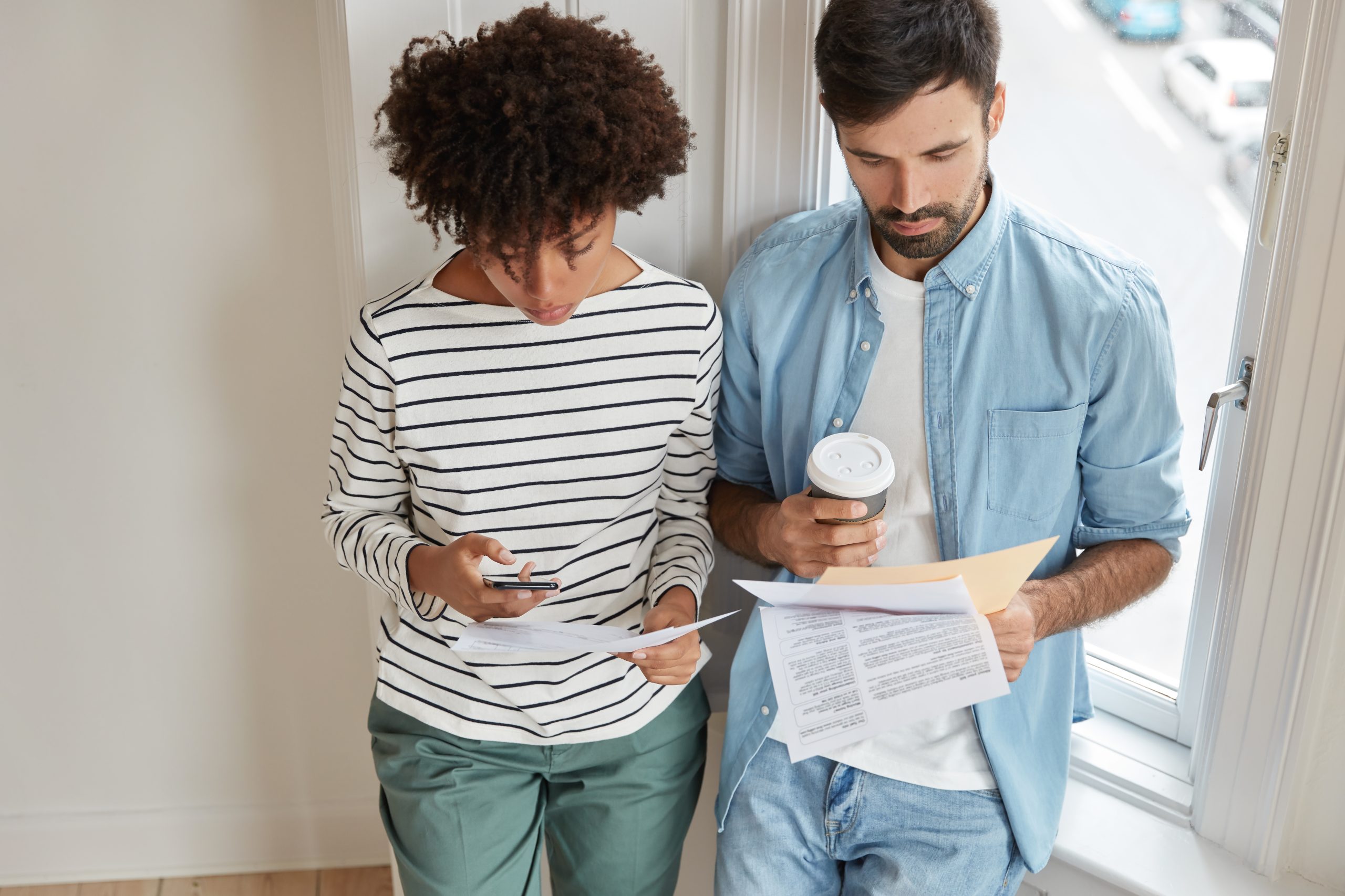 Top view of business coach has informal meeting with trainee, tries explain some information, holds papers and takeaway coffee, dark skinned woman checks something on cell phone, stand at window sill