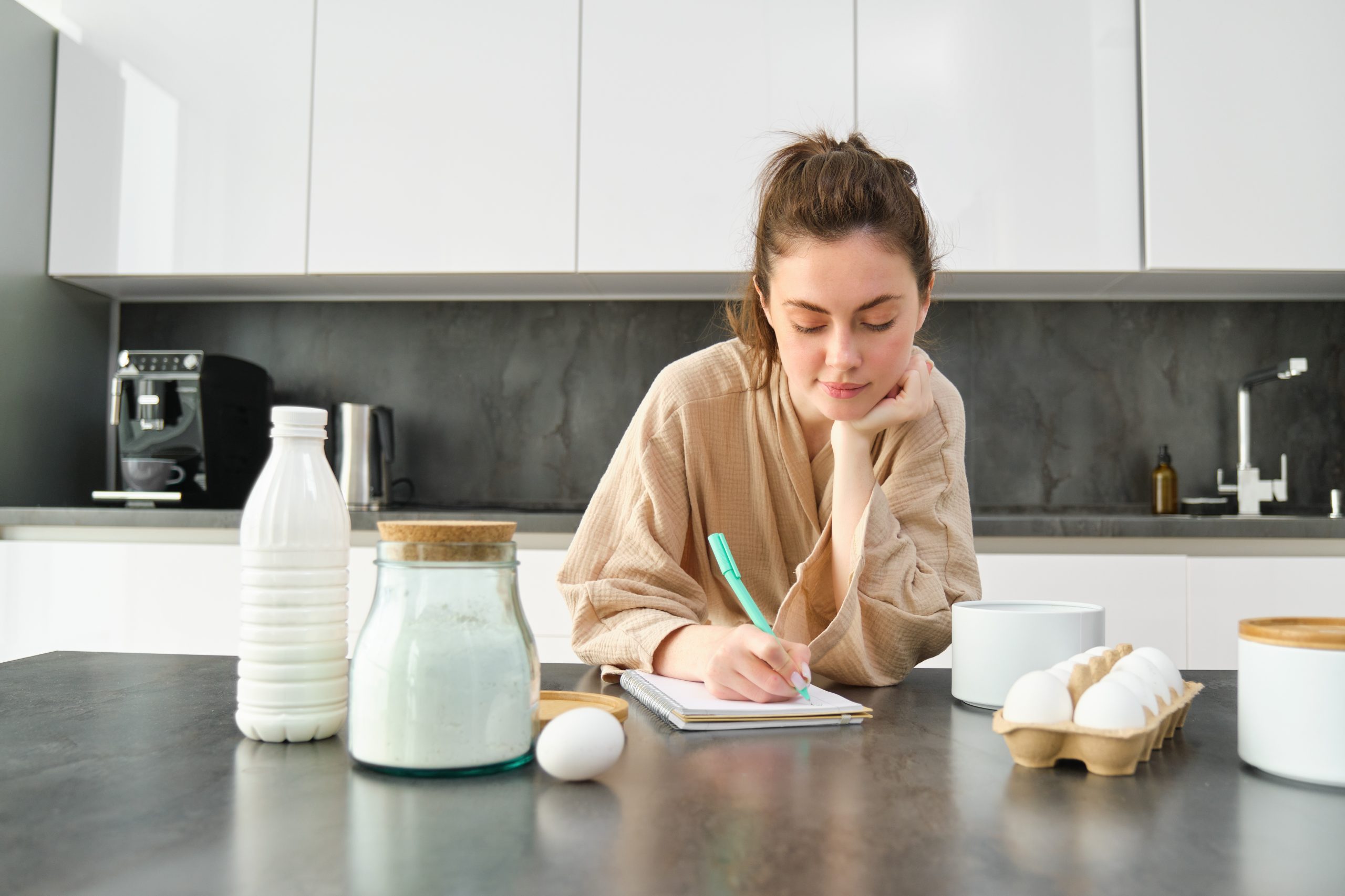 Attractive young cheerful girl baking at the kitchen, making dough, holding recipe book, having ideas